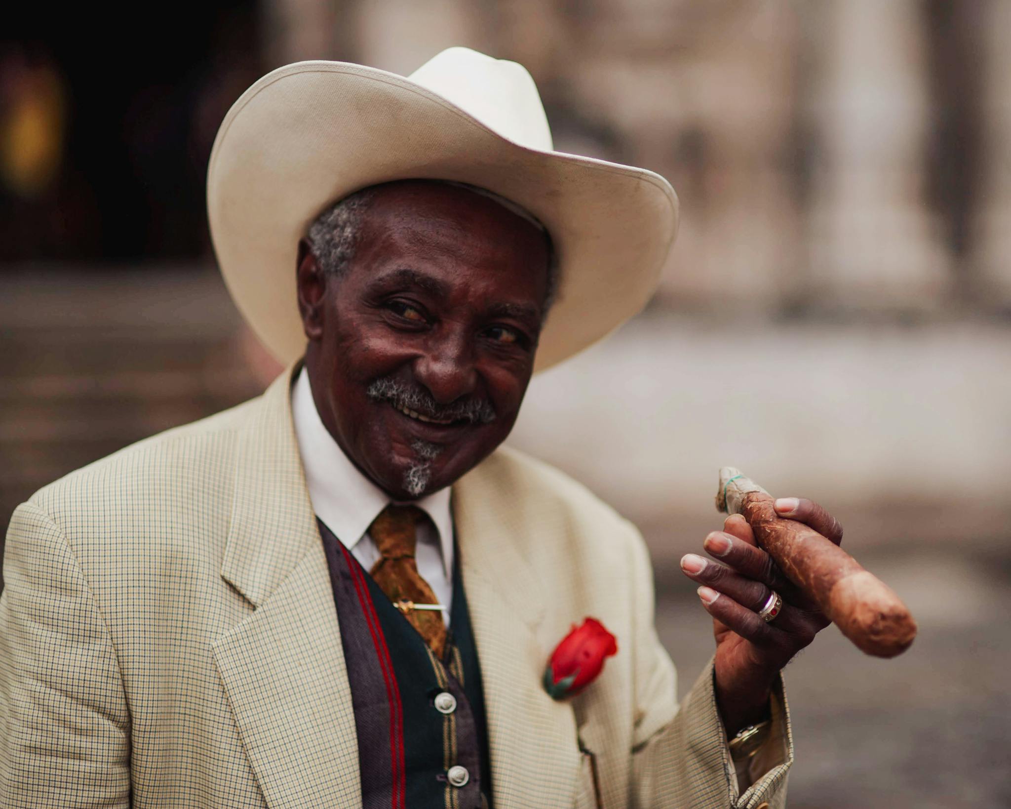 A senior man in a white hat and suit smiling while holding a cigar.