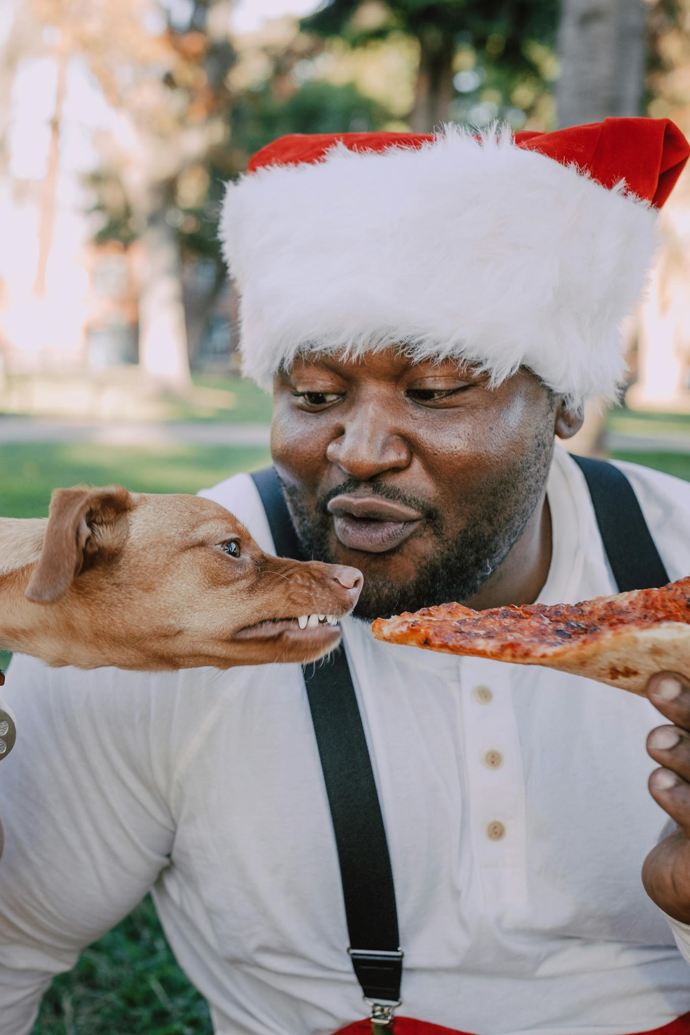 Cheerful man in Santa hat sharing a slice of pizza with a dog in a park setting.