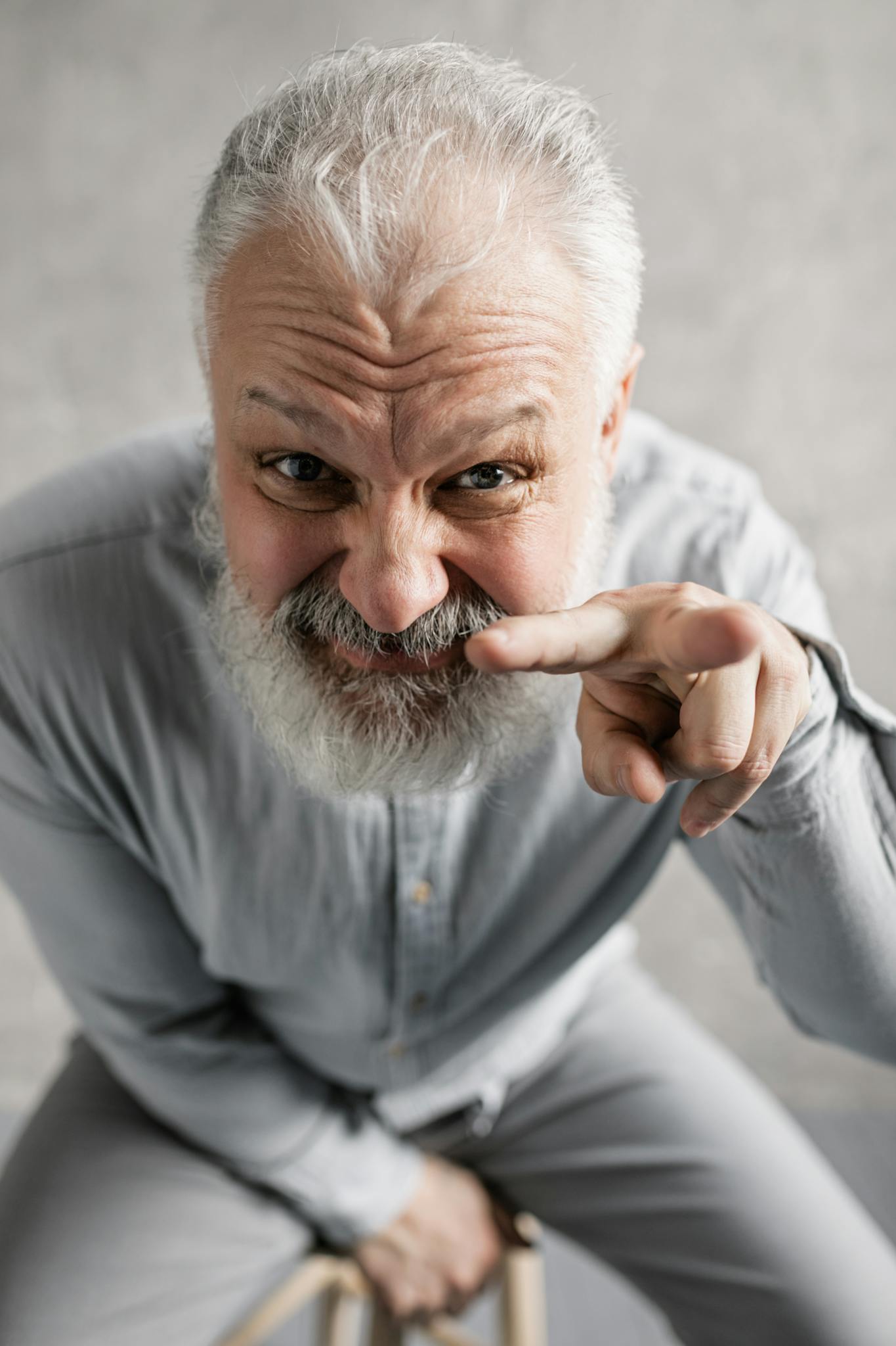 Elderly man with gray hair and beard smiling playfully indoors, wearing gray shirt.